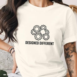 Puede incluir: Camiseta color crema con un diseño de engranajes negros y el texto "DESIGNED DIFFERENT". El gráfico incluye engranajes entrelazados, transmitiendo un tema de innovación e ingeniería. La camiseta es de material suave.