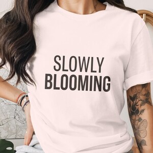 Może przedstawiać: Jasnoróżowy t-shirt z krótkim rękawem z napisem SLOWLY BLOOMING nadrukowanym pogrubionymi czarnymi wielkimi literami na środku klatki piersiowej. Odzież posiada klasyczny okrągły dekolt oraz proste wykończenie dołu, co zapewnia prosty krój.