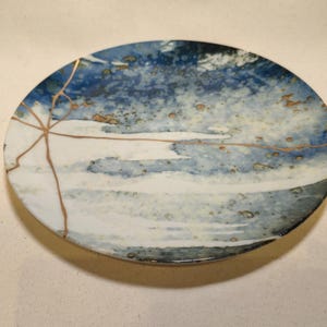 May include: A round ceramic plate featuring a mottled blue and white glaze with a textured, speckled finish. The surface is accented by thin, branching gold-colored lines that traverse the plate, creating a distinct pattern across the surface.