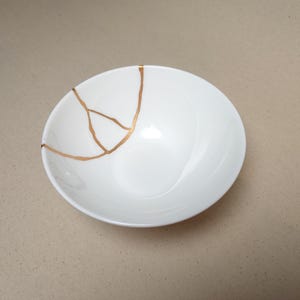 Könnte beinhalten: Eine weiße Keramikschale mit einer Reparaturstelle in metallischer Goldfarbe. Die Schale hat ein glattes, abgerundetes Inneres und einen geschwungenen Rand, wobei sich die goldfarbene Reparatur vom Rand bis zur Mitte des Beckens erstreckt.
