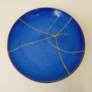 May include: A round ceramic plate in a deep cobalt blue glaze featuring prominent gold-colored lines that traverse the surface in a pattern resembling repaired cracks, with a dark brown rim visible along the outer edge of the circular dish.