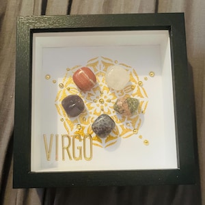Puede incluir: Una exhibición enmarcada con cinco piedras preciosas pulidas dispuestas sobre un diseño de mandala dorado. La palabra "VIRGO" se muestra en letras doradas. El marco es negro.
