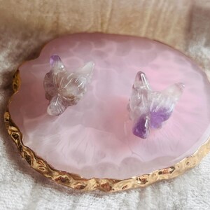 May include: Two carved amethyst crystal bird figurines sit on a pink agate slice with a gold-coloured edge. The birds are purple and clear crystal. The agate slice is a decorative item.