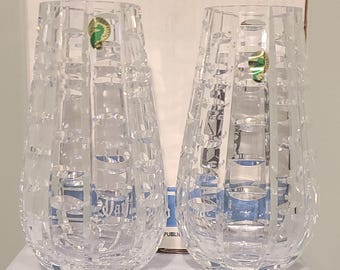 Puede incluir: Dos jarrones de cristal transparente con un diseño geométrico texturizado. Cada jarrón tiene forma de lágrima y una pegatina verde y dorada. Los jarrones se exhiben frente a una caja blanca.