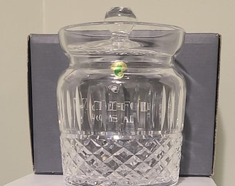 May include: A clear glass jar with a lid, featuring a diamond-cut pattern on the lower half and vertical lines on the upper half. The jar has a round shape and a clear glass knob on top. The word "WATERFORD" is etched on the front.