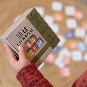 Könnte beinhalten: Eine beige Box des Elif Ba Lernspiel-Spiels mit bunten quadratischen Kacheln mit arabischen Buchstaben. Die Box hat Text auf Deutsch und Arabisch. Das Spiel ist für spielerisches Lernen, Feinmotorik und Konzentration konzipiert.