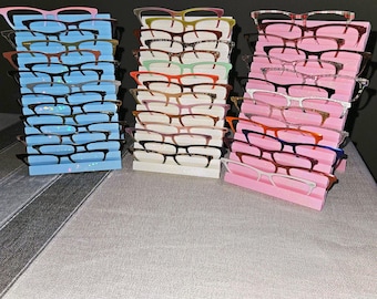 May include: Three stacks of eyeglasses in various colors, including pink, blue, and white, are displayed on a light-colored surface. Each stack holds multiple pairs of glasses, showcasing a variety of frame styles and colors.