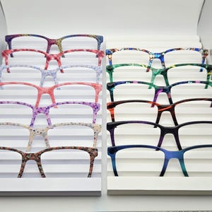 May include: A collection of twelve pairs of spectacles with various patterned and solid coloured frames arranged in two vertical columns on a white tiered display stand. The frames feature diverse designs including floral, speckled, and gradient patterns.