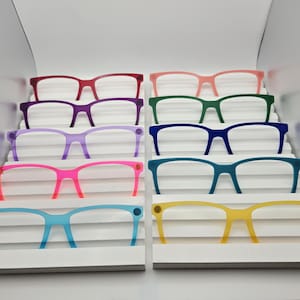 May include: A collection of ten pairs of plastic reading glasses arranged in two columns on a white tiered display stand. The frames feature a variety of solid colors including red, purple, light purple, hot pink, light blue, peach, green, dark blue, teal, and yellow.