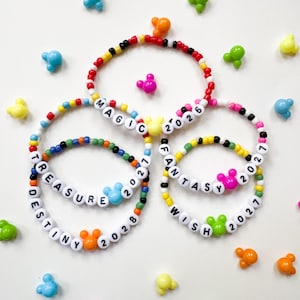 May include: Four circular bracelets made of colorful plastic beads. The bracelets feature white letter beads spelling MAGIC 2026, TREASURE 2027, DESTINY 2028, and FANTASY 2027. Each bracelet includes a small, colorful, mouse-ear-shaped plastic charm.