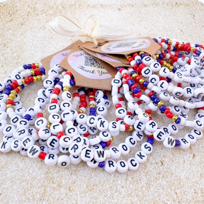 Pu&ograve; includere: Una collezione di braccialetti di perline bianche con perline di lettere che compongono "CREW ROCKS". I braccialetti presentano perline di accento rosse, blu e dorate. I braccialetti sono legati con un nastro ed etichette che dicono "Grazie".