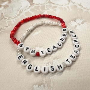 Peut inclure: Deux bracelets de perles. L'un est rouge et blanc, l'autre est blanc avec des lettres noires &eacute;pelant "ENGLISH TEACHER" et "GYM TEACHER". Les bracelets sont sur une surface en dentelle blanche.