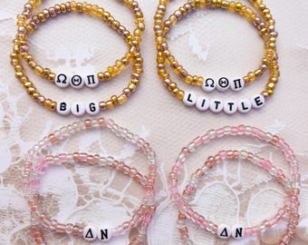 May include: Four beaded bracelets arranged in pairs. The top two are gold-toned beads with white letter beads spelling BIG and LITTLE with Greek letters. The bottom two are pink-toned beads with white letter beads spelling BIG and LITTLE with Greek letters.
