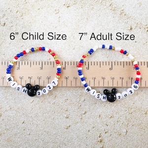 K&ouml;nnte beinhalten: Zwei Armb&auml;nder mit Perlen, eines in Kindergr&ouml;&szlig;e (15 cm) und eines in Erwachsenengr&ouml;&szlig;e (18 cm). Die Armb&auml;nder haben rote, wei&szlig;e, blaue und goldene Perlen, einen schwarzen Mickey Mouse Kopf und die Worte "WISH 2021" und "WISH 2026".