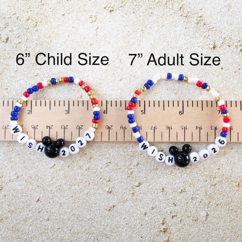 K&ouml;nnte beinhalten: Zwei Armb&auml;nder mit Perlen, eines in Kindergr&ouml;&szlig;e (15 cm) und eines in Erwachsenengr&ouml;&szlig;e (18 cm). Die Armb&auml;nder haben rote, wei&szlig;e, blaue und goldene Perlen, einen schwarzen Mickey Mouse Kopf und die Worte "WISH 2021" und "WISH 2026".