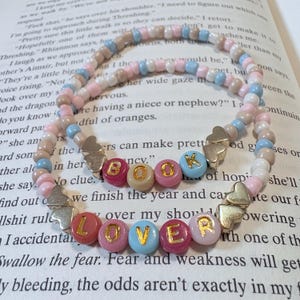 May include: Two beaded bracelets with the words "BOOK" and "LOVER" spelled out in colorful letter beads. The bracelets feature pink, blue, and tan beads, with silver heart and butterfly charms. The bracelets are on a book page.