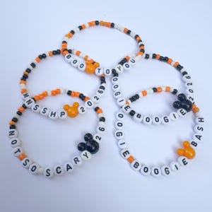 May include: A collection of five circular beaded bracelets featuring orange, black, and white seed beads. The bracelets include white letter beads spelling out phrases like BOO TO YOU, MNSSHP 2026, NOT SO SCARY 26, and OOGIE BOOGIE BASH, with orange or black mouse-shaped beads.