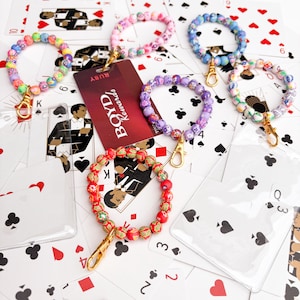 May include: A collection of beaded wristlet keychains in various colors, including red, purple, pink, and blue, with gold-colored clasps. The keychains are displayed on a background of playing cards and a red rewards card.
