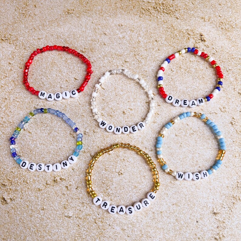Pu&ograve; includere: Sei braccialetti di perline con perline di lettere bianche che compongono le parole "MAGIC", "WONDER", "DREAM", "DESTINY", "TREASURE" e "WISH". I braccialetti presentano perline rosse, bianche, blu e dorate. I braccialetti sono su una superficie color sabbia.