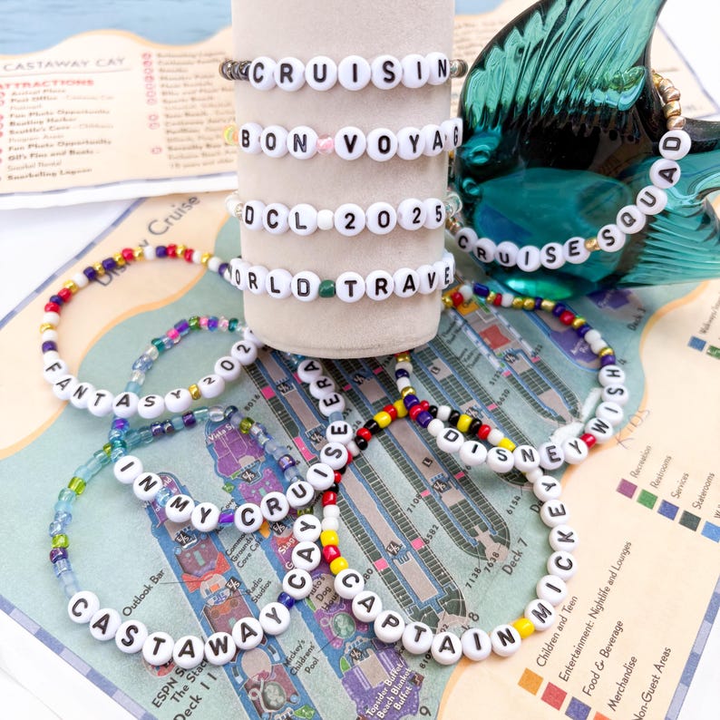 Pu&ograve; includere: Una collezione di braccialetti di perline con perline di lettere bianche che compongono frasi come "Cruisin", "Bon Voya", "DCL 2025" e "World Travel". Altri braccialetti dicono "Castaway Cay", "In My Cruise Era" e "Disney Wish". I braccialetti sono su una mappa.