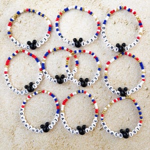 May include: A collection of beaded bracelets with red, white, blue, and gold beads. Each bracelet features a black Mickey Mouse head charm and white letter beads spelling out words like "Magic," "Wish," and "Adventure," along with the year 2026 or 2027.