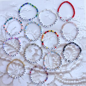 Peut inclure: Plusieurs bracelets de perles avec des lettres blanches &eacute;pelant des phrases comme "Engaged Era", "Swiftie" et "All Too Wed". Les bracelets pr&eacute;sentent diff&eacute;rentes couleurs de perles, dont le bleu, le rose et le rouge, et sont pr&eacute;sent&eacute;s sur une surface en dentelle blanche.