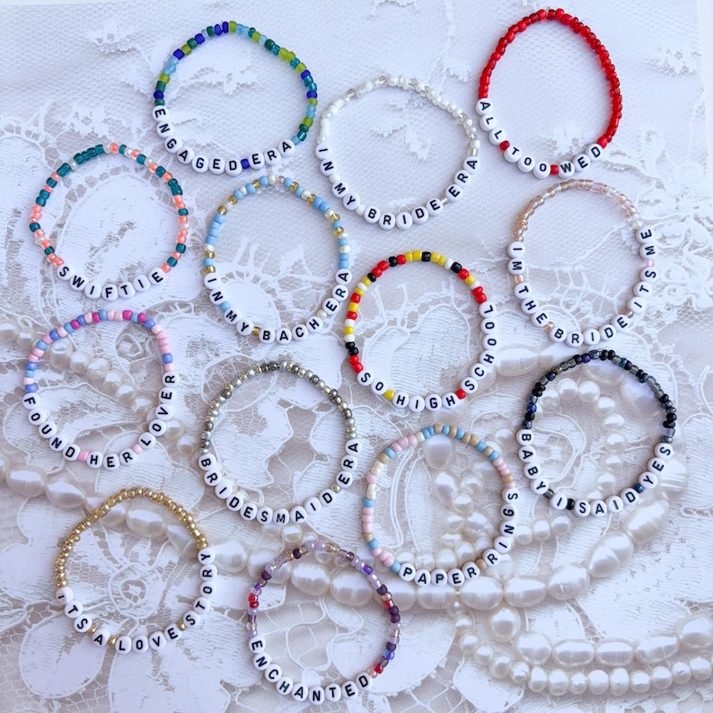 Peut inclure: Plusieurs bracelets de perles avec des lettres blanches &eacute;pelant des phrases comme "Engaged Era", "Swiftie" et "All Too Wed". Les bracelets pr&eacute;sentent diff&eacute;rentes couleurs de perles, dont le bleu, le rose et le rouge, et sont pr&eacute;sent&eacute;s sur une surface en dentelle blanche.