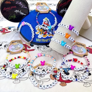 May include: A collection of colorful beaded bracelets with white letter beads spelling out phrases like "CAST MEMBER" and "MAKE MAGIC." The bracelets feature Mickey Mouse head charms and are displayed with a blue hat and a decorative stand. Each bracelet has a tag that says "Thank you for making magic."