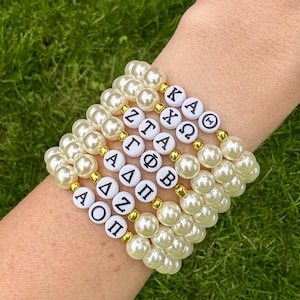 May include: A stack of pearl bracelets with gold accents and white letter beads. The bracelets feature Greek letters, including Alpha, Omega, and Pi. The bracelets are on a wrist against a green background.