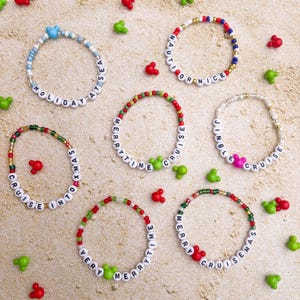 May include: A collection of beaded bracelets with white letter beads spelling out phrases like "Holiday at Sea" and "Merrytime Cruise." The bracelets feature colorful beads and Mickey Mouse-shaped accents in red, green, and blue.