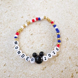 K&ouml;nnte beinhalten: Ein Perlenarmband mit wei&szlig;en Buchstabenperlen, die "WONDER 2026" buchstabieren, und einem schwarzen Mickey Mouse Kopf-Charm. Das Armband hat rote, wei&szlig;e, blaue und goldene Perlen. Das Armband liegt auf einer hellbraunen Oberfl&auml;che.
