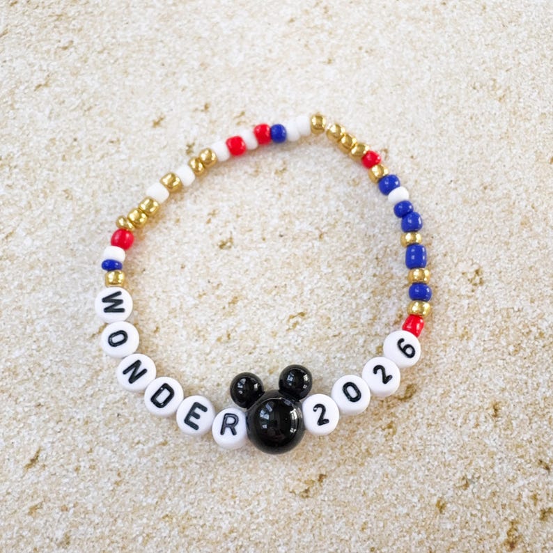 K&ouml;nnte beinhalten: Ein Perlenarmband mit wei&szlig;en Buchstabenperlen, die "WONDER 2026" buchstabieren, und einem schwarzen Mickey Mouse Kopf-Charm. Das Armband hat rote, wei&szlig;e, blaue und goldene Perlen. Das Armband liegt auf einer hellbraunen Oberfl&auml;che.