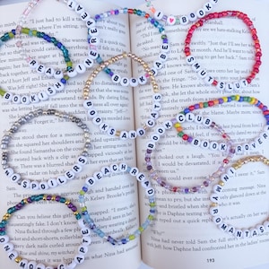 May include: White beaded bracelets with black letter beads spelling out words like "BOOKS", "SPOILERS", and "BEACH READ". The bracelets are on an open book with text. Colorful beads accent some bracelets.