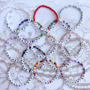 Peut inclure: Plusieurs bracelets de perles avec des perles de lettres blanches &eacute;pelant des phrases comme "In My Bride Era" et "All Too Wed". Les bracelets pr&eacute;sentent diverses couleurs, dont le rouge, le bleu et le rose, et sont pr&eacute;sent&eacute;s sur une surface en dentelle blanche.