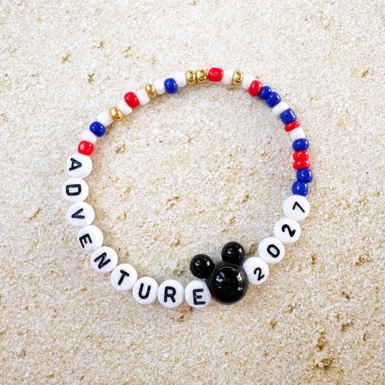 K&ouml;nnte beinhalten: Ein Perlenarmband mit wei&szlig;en, roten, blauen und goldenen Perlen. Das Armband schreibt "ADVENTURE 2021" mit wei&szlig;en Buchstabenperlen. Ein schwarzer Mickey Mouse Kopf-Charm ist ebenfalls enthalten.