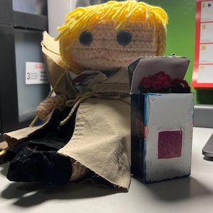 May include: A crocheted doll with yellow yarn hair, black button eyes, and a beige trench coat over a black outfit. Beside the doll is a small white rectangular box with a red square on the front and a red crocheted item inside the open top.