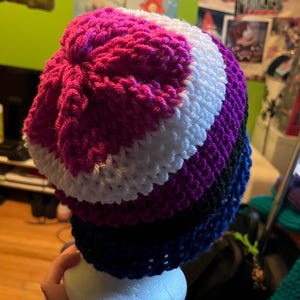 May include: A handmade crochet beanie in a bi-pride color scheme. The hat features stripes of pink, white, purple, black, and blue. The hat is displayed on a white foam head.