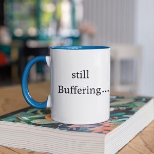 May include: White ceramic mug with a blue interior and handle. The mug has the words "still Buffering..." printed in black. The mug is sitting on a book with a colorful floral design.