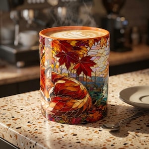 May include: A cylindrical ceramic mug featuring a stained-glass style illustration of a curled-up fox surrounded by vibrant autumn maple leaves in shades of red, orange, and gold. The mug contains a hot beverage with latte art on the surface.