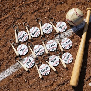 May include: A baseball-themed collection featuring personalized keychains, a baseball, and a wooden bat. The keychains are designed as baseballs with names and numbers. The baseball and bat are positioned on a baseball field.