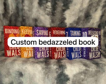 May include: A collection of custom bedazzled books in various colors, including red, orange, purple, and blue. Each book has a title and author's name, with the text "Custom bedazzled book" overlaid on the image.