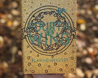 May include: A gold-colored book cover with a textured surface and a circular design in black, teal, and gold. The text "Fourth Wing" is visible within the design. The book is held in a hand.