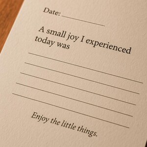 May include: Close-up of a cream-colored journal page with the text "Date:" and lines for writing. The text "A small joy I experienced today was" is printed above the lines, and "Enjoy the little things." is printed at the bottom.