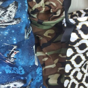 May include: Three fabric bags hanging from the headrests of a car. The bags are made of different fabrics, one with a camouflage pattern, one with a black and white geometric pattern, and one with a blue background and a white spaceship pattern with the words "Star Wars" printed on it.