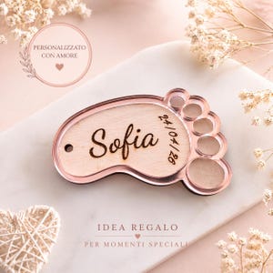 May include: A personalized wooden baby footprint keepsake with the name "Sofia" and the date 24/04/26 engraved on it. The footprint is framed in a rose gold color. The text "PERSONALIZZATO CON AMORE" is in a circle.