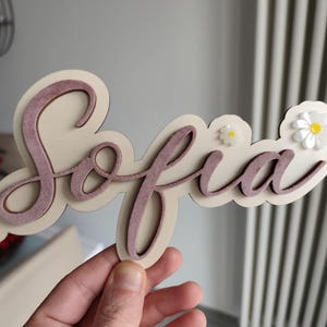Può includere: Un cartello in legno con il nome "Sofia" in corsivo, sovrapposto a una base a forma di nuvola beige chiaro. Le lettere sono di colore malva e il cartello è decorato con piccoli fiori di margherita bianchi.