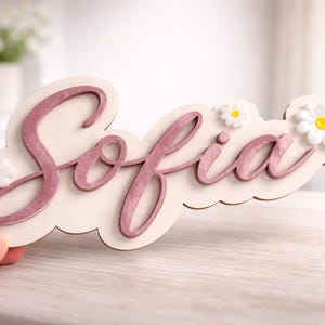 Puede incluir: Un letrero de nombre de madera que dice "Sofia" en una fuente cursiva, de color rosa-púrpura. El letrero está decorado con pequeñas margaritas blancas con centros amarillos. El letrero está sostenido por una mano.