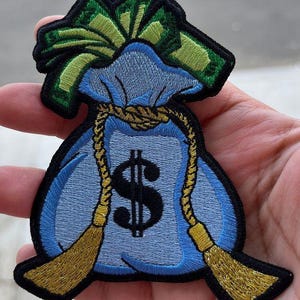 Może przedstawiać: Haftowana naszywka przedstawiająca jasnoniebieski worek na pieniądze z czarnym znakiem dolara. Worek jest zawiązany złotą liną i ma zielone i ż&oacute;łte akcenty. Naszywka ma czarną obw&oacute;dkę.