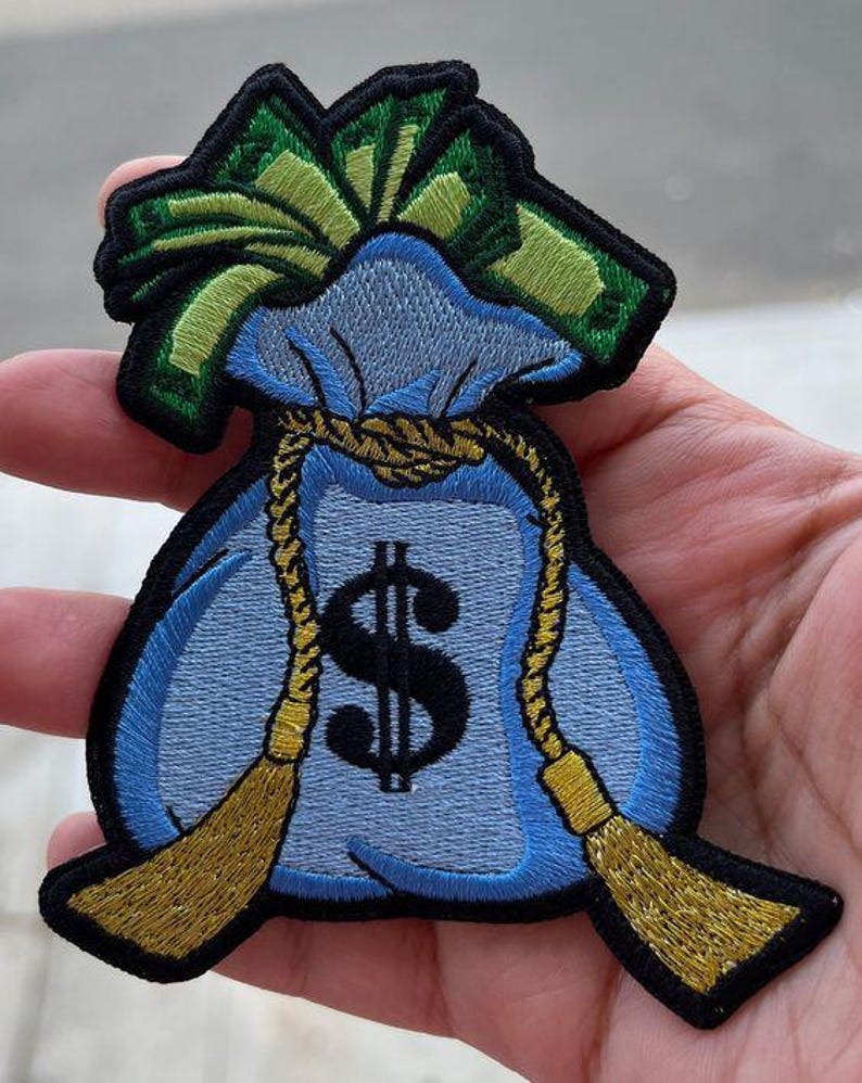 Może przedstawiać: Haftowana naszywka przedstawiająca jasnoniebieski worek na pieniądze z czarnym znakiem dolara. Worek jest zawiązany złotą liną i ma zielone i ż&oacute;łte akcenty. Naszywka ma czarną obw&oacute;dkę.