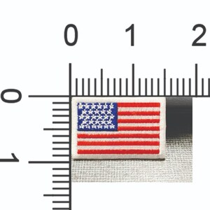 Może przedstawiać: Haftowana naszywka z flagą amerykańską. Naszywka ma białą obw&oacute;dkę, niebieskie pole z białymi gwiazdami oraz czerwono-białe paski. Naszywka ma wymiary około 2,5 cm na 1,3 cm.